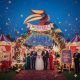 "A grand carnival-themed wedding entrance featuring a large colorful hornbill sculpture above a floral archway. The sign reads Hornbill Event Planner, with a bride, groom, and wedding party standing on a red carpet amidst confetti, circus tents, and vintage popcorn machines